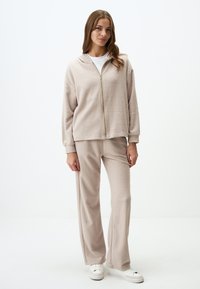 Felpa beige con zip davanti in morbido tessuto, maniche lunghe, abbinata a pantaloni a gamba larga coordinati. Scarpe da ginnastica bianche completano l'outfit.