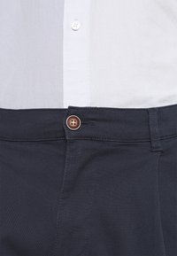 Mörkblå byxor med en slät yta, med en brun knapp i midjan och vit skjorttyg synligt vid den övre kanten.