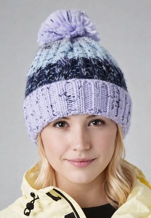 Young woman with light blonde hair wears a lilac, blue, and black knitted beanie with a pom-pom and a pale yellow jacket with a zipper.