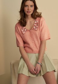 Kurzärmlige, korallenrosa Strickjacke mit floraler Stickerei am Kragen, Knopfverschluss, kombiniert mit einem plissierten hellgrünen Rock.