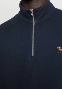 Marine blauwe ritspullover met hoge kraag, voorzien van een zilveren rits en een kleine, oranje hert-embroidery op de linkerborst. Van een katoenmix.