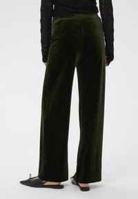 Pantaloni largi din catifea verde închis cu o textură netedă. Aceștia au un talie înaltă și o croială lejeră, completată de pantofi negri tip slip-on.
