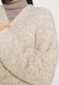 Béžový huňatý pletený svetr na knoflíky s výstřihem do V a nadměrnými rukávy. Viditelná texturovaná látka, nošený přes hnědý rolák.