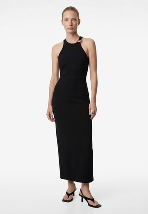 Donna che indossa un vestito nero senza maniche fino alla caviglia con collo allacciato al collo e dettaglio con fibbia, abbinato a sandali neri con tacco e anelli.