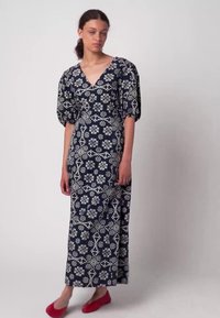 Granatowa sukienka maxi z białym kwiatowym wzorem, głębokim dekoltem w serek i bufkowymi rękawami. Modelka ma na sobie czerwone baleriny, prezentując pełną długość sukienki.