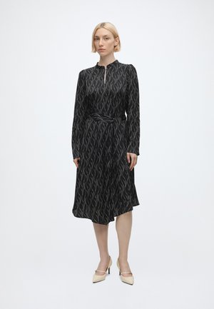 Femme blonde debout portant une robe midi noire à manches longues avec un motif ondulé argenté et des chaussures à talons beiges sur fond blanc.