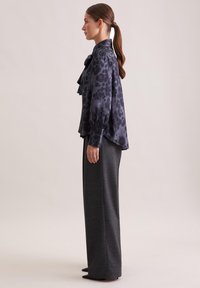 Blusa di seta di un blu navy scuro con motivo a leopardo, caratterizzata da un colletto arricciato e maniche lunghe; abbinata a pantaloni grigi a vita alta.