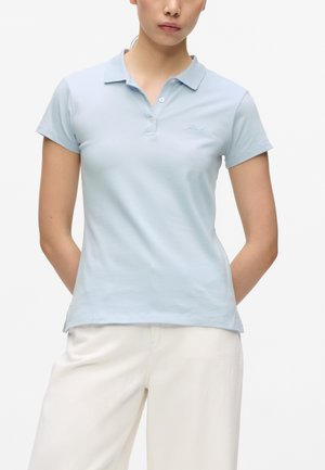 Kvinde iført en lyseblå kortærmet polo og offwhite bukser med hænderne bag ryggen mod en hvid baggrund.