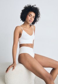 Witte geribbelde bralette en hooggesneden onderbroek van gladde stof, met verstelbare bandjes, tegen een neutrale achtergrond.