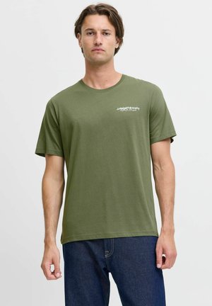 Hombre con camiseta de manga corta color verde oliva con pequeño logo blanco en el pecho y vaqueros azul oscuro, de pie frente a un fondo liso y claro.