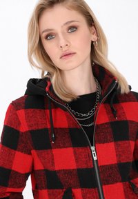 Felpa con zip a quadri rossi e neri, con cappuccio nero, dotata di chiusura con zip e lacci. Il modello indossa catene argento sovrapposte.