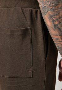 Braune texturierte Hose mit einer Seitentasche und subtiler Dreiecksakzentuierung. Der Stoff wirkt strapazierfähig und strukturiert. Nahaufnahme des Materials.
