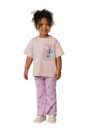 Göndör hajú kisgyerek rózsaszín pólóban, amelyen rajzfilmfigura látható, és lila virágmintás nadrágban, mosolyogva és fehér háttér előtt állva.