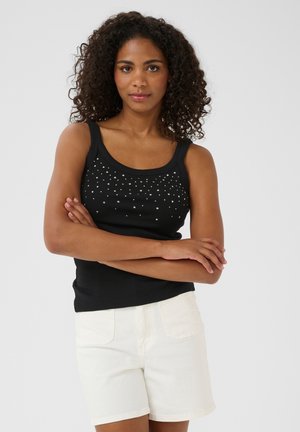 Top negro sin mangas con textura acanalada, que presenta tachuelas plateadas dispuestas en un patrón en la parte frontal. Combinado con pantalones cortos de mezclilla blancos.