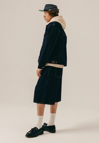 Person trägt eine dunkle Jeansjacke und knielange Shorts, einen cremefarbenen Hoodie, eine gestreifte Mütze, weiße Socken und schwarze Tassel-Loafer, steht seitlich.