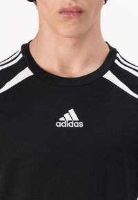 T-shirt de sport noir avec des accents blancs et trois bandes sur les épaules. Présente un logo Adidas blanc sur la zone de la poitrine. Tissu doux.