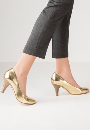 Des escarpins en cuir verni doré avec un bout rond, un talon strié de 10 cm et une finition lisse et brillante. Portés avec des pantalons gris à la coupe droite.