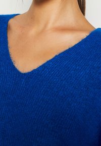 Blå stickad tröja med V-ringning, med ett strukturerat, mjukt tyg och en lös passform. Närbild som betonar halsringningen och strukturen.