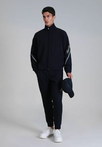Navyblauer Zip-Up-Jacke mit weißen Akzenten, kombiniert mit passenden Jogginghosen. Das Modell hält eine schwarze Mütze. Weiße Sneakers runden den Look ab.