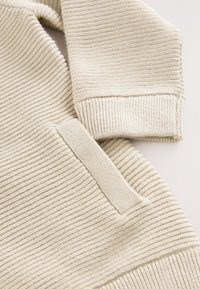 Pull beige texturé avec tissu côtelé, une poche appliquée et des poignets ajustés. Le matériau semble tricoté et doux avec un design contemporain.
