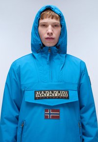 Giacca sintetica blu con cappuccio, con una tasca frontale, patch con logo e dettaglio della bandiera norvegese. Texture liscia e design minimal.