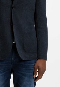 Granatowa marynarka w jodełkę, dwuguzikowa, z usztywnionymi ramionami i naszywanymi kieszeniami, zestawiona z ciemnoniebieskimi dżinsami.