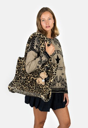 Leopard print tote bag made of soft, textured fabric. Features a plush teddy bear keychain and rounded edges. Model wears a patterned sweater.