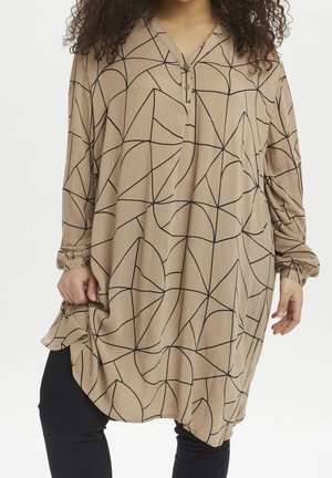 Kobieta ubrana w luźną beżową tunikę z czarnymi geometrycznymi liniami na czarnych spodniach, ręce poprawiające dół i rękaw tuniki.