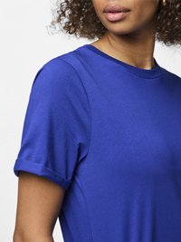 Kvinna som bär en kungligt blå t-shirt med korta ärmar och uppvikta fållar, lockigt hår och neutralt läppstift mot en enkel vit bakgrund.