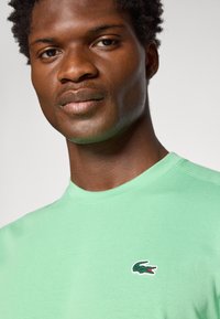 Detailný záber muža s tmavou pokožkou v svetlozelenej košeli s malým logom krokodíla na hrudi na jednofarebnom pozadí.