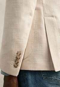 Béžová blazer s textúrovanou tkaninou, so zapínaním na tri gombíky a bočným rozparkom. V kombinácii s džínovou košou.