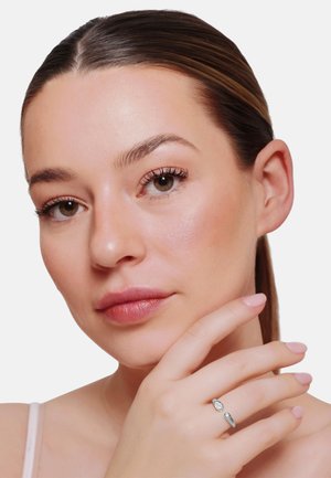 Vrouw met een gladde huid en glanzend haar raakt haar kin aan en toont een zilveren ring met twee heldere stenen aan haar vinger.