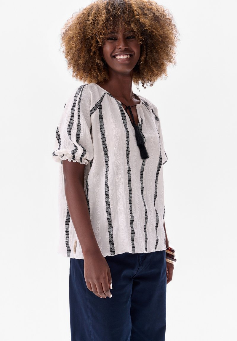 Witte blouse met zwarte verticale strepen, lichte textuurstof, losse pasvorm, geplooide mouwen en een kwastje detail bij de halslijn.
