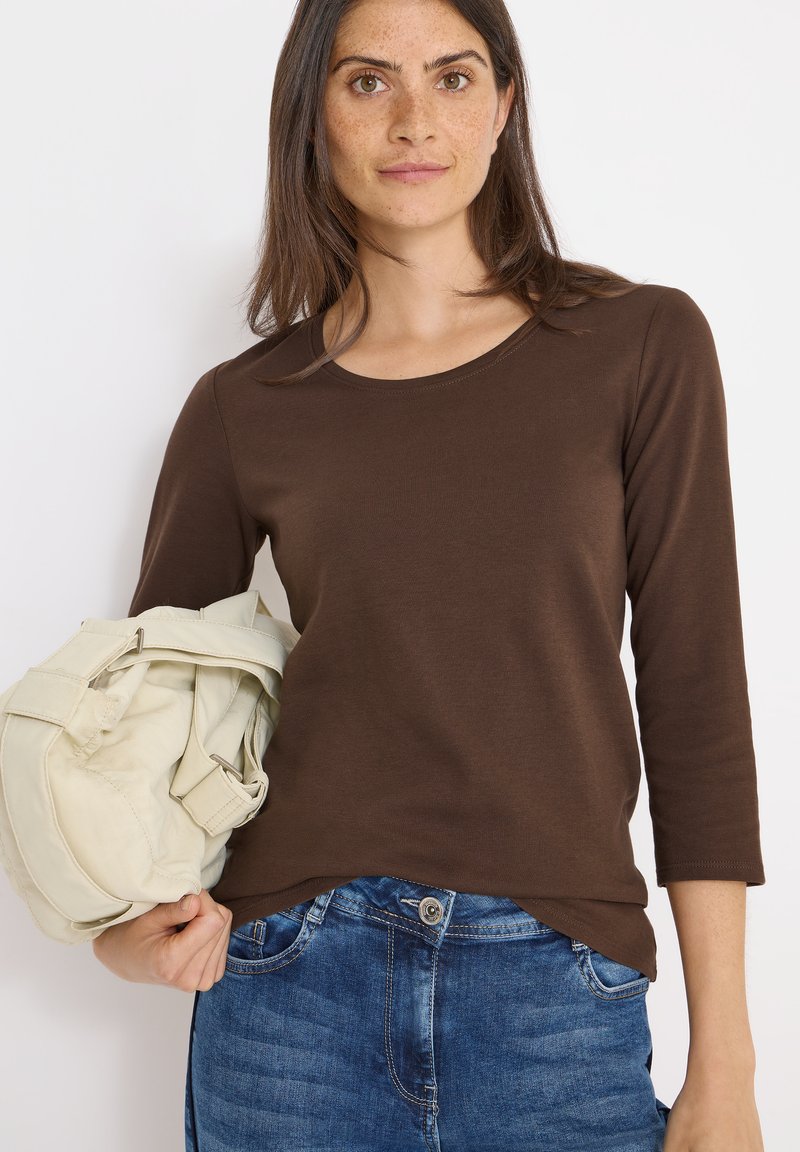 Maglia marrone a maniche lunghe con scollo rotondo, abbinata a jeans blu. La modella tiene una borsa chiara con una superficie texture.