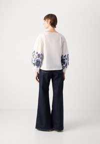 Weiße Bluse mit blauer floralem Stickerei an den Ärmeln, kombiniert mit dunkelblauen, ausladenden Jeans. Glatte Textur und lockerer Schnitt.