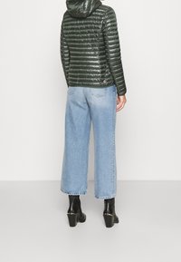 Tmavě zelená péřová bunda s kapucí, s horizontálním prošíváním a logem. V kombinaci s světle modrými širokými džíny a černými koženými botami na podpatku.