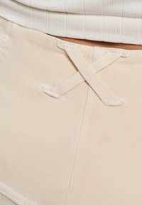 Nærbilde av beige bukser med en liten sydd stoff "X" detalj nær midjebåndet under en hvit ribbestrikket skjorte.