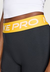 A Nike Pro fekete sportlegging sárga gumírozott derékkal rendelkezik, melyen fehér "NIKE PRO" felirat található. Az anyag sima, rugalmas textúrájú.