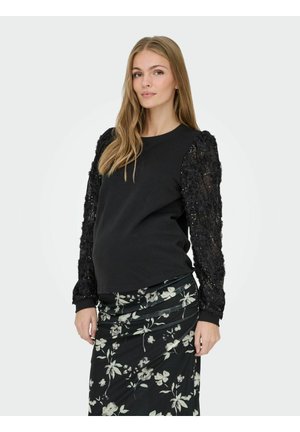 Sweat-shirt noir avec des manches texturées et pailletées ; le modèle le porte avec une jupe noire à imprimé floral, dotée d'un design froncé.