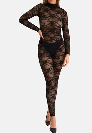 Kobieta w czarnym, całkowicie przezroczystym koronkowym body z długimi rękawami i wysokim kołnierzem, w połączeniu z czarnymi wysokimi obcasami, stojąca z jedną ręką na biodrze.