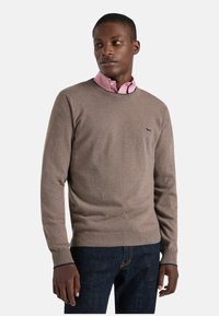 Maglione marrone in tessuto morbido, con scollo rotondo, polsini a costine e un piccolo logo ricamato sul petto, indossato sopra una camicia rosa.