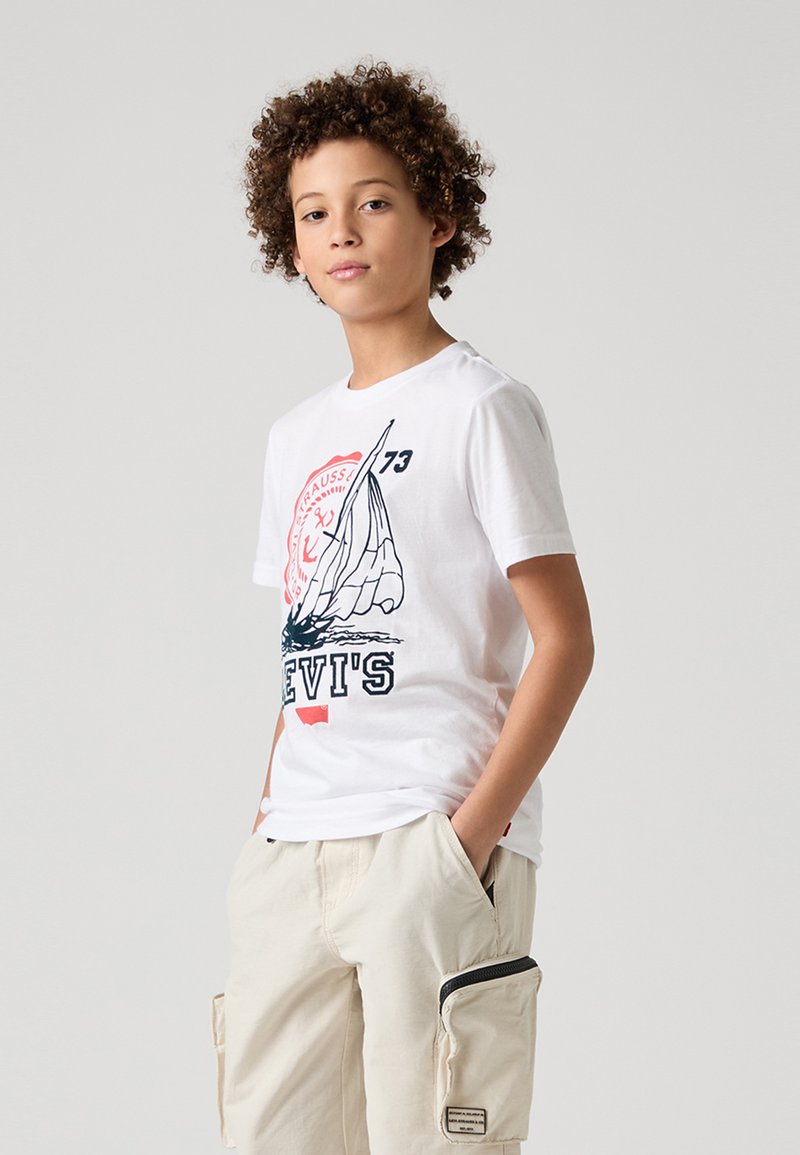 Camiseta blanca de algodón con un diseño gráfico de velero, con detalles en rojo y azul marino. Combinada con pantalones cortos de cargo en beige claro que exhiben bolsillos.