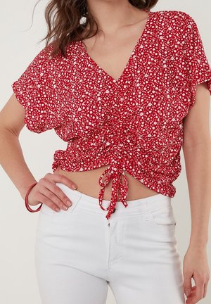 Woman wearing a red floral short-sleeve blouse with drawstring front and white high-waist jeans, hand on hip with matching bracelet.