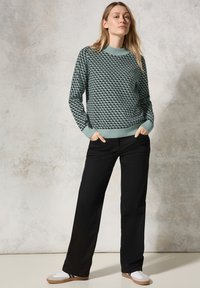 Groene gebreide trui met geometrisch patroon, ronde halslijn en geribbelde manchetten, gecombineerd met zwarte broek met wijde pijpen en witte sneakers.