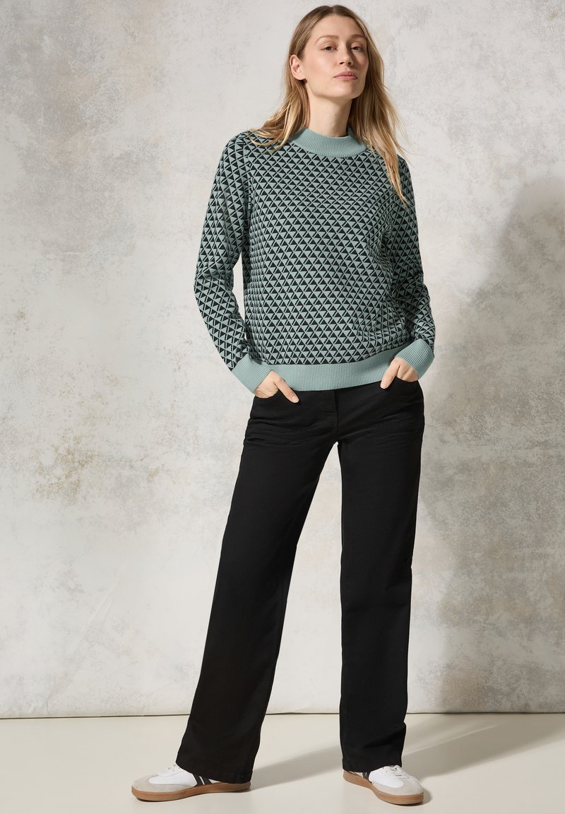 Groene gebreide trui met geometrisch patroon, ronde halslijn en geribbelde manchetten, gecombineerd met zwarte broek met wijde pijpen en witte sneakers.