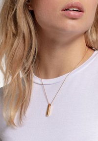 Collana con ciondolo in oro a forma rettangolare su una sottile catena, indossata sopra una camicia bianca, con una texture metallica liscia.