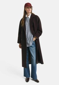 Manteau long en laine bordeaux foncé porté sur une chemise à rayures bleues et blanches, associé à un jean évasé et des bottes marron foncé. Casquette bordeaux avec un logo.