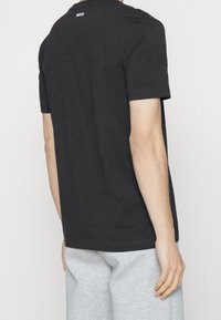 Person klädd i en svart t-shirt med kort ärm och "BOSS"-logotyp på övre ryggen, tillsammans med ljusgrå byxor mot en neutral bakgrund.