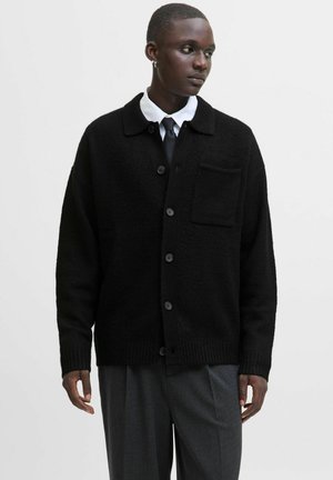 Black, textured cardigan with a collar, front button closure, and a chest pocket. Worn over a white shirt and tie, paired with gray trousers.