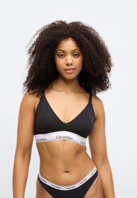 Crni pamučni bralette s dizajnom u obliku slova V. Ima bijelu elastičnu traku s logom "Calvin Klein". Podesive naramenice za prilagodbu.